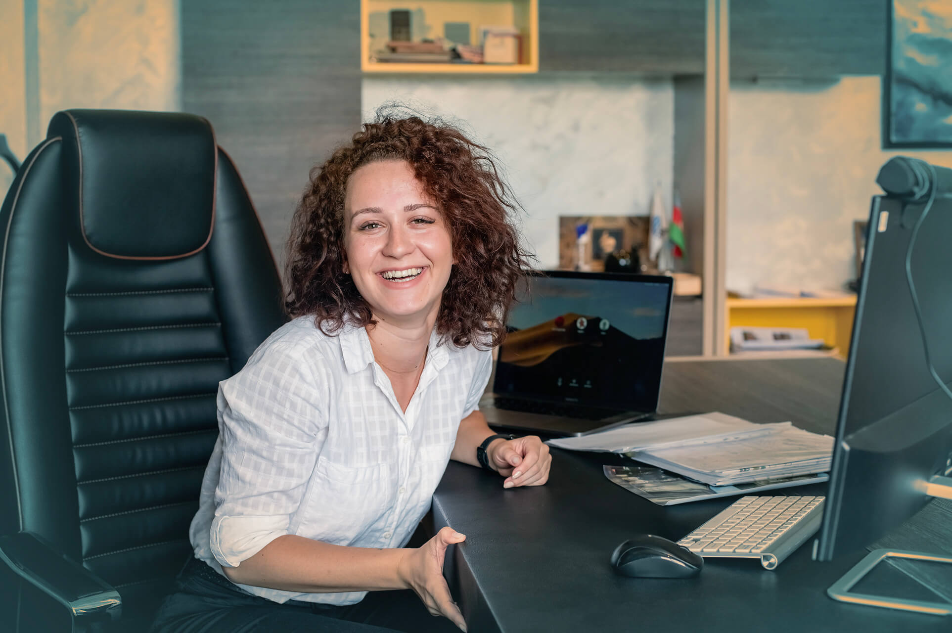 A woman with curly hair smiling while sitting at a desk with a computer, papers, and a laptop in a modern office.