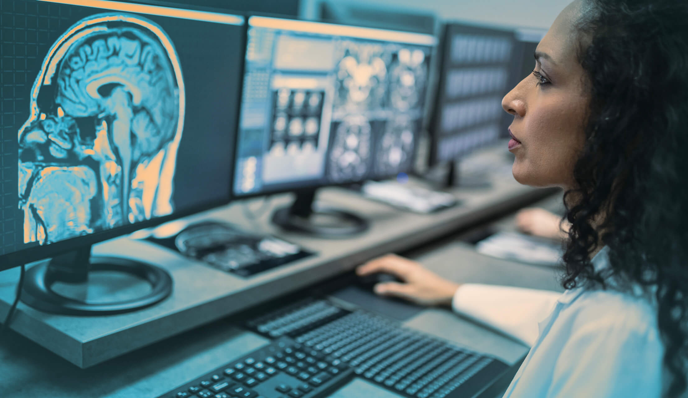 A woman analyzes brain scan images on multiple computer monitors in a cybersecurity research lab.