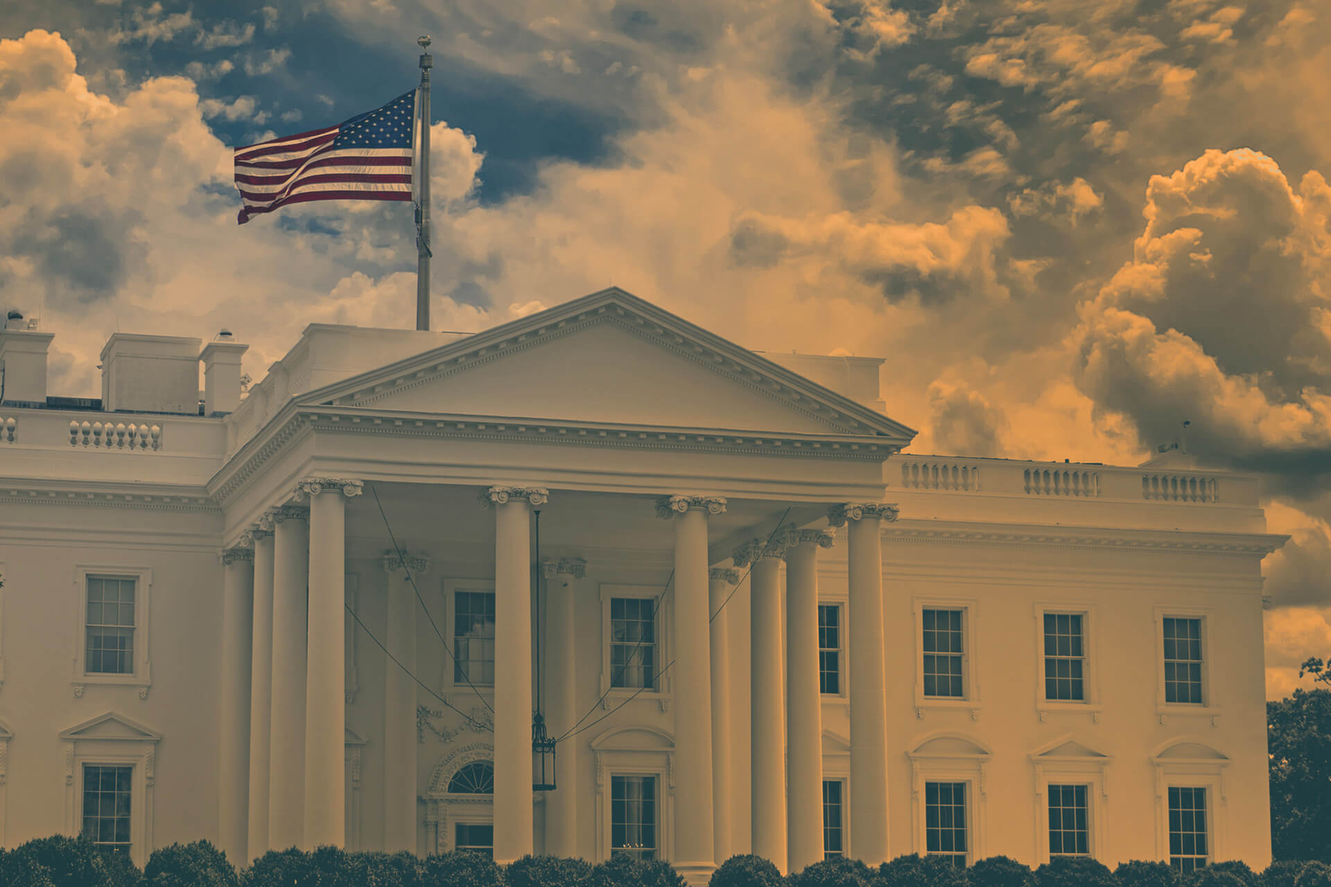 The White House with an American flag flying against a cloudy sky at sunset.