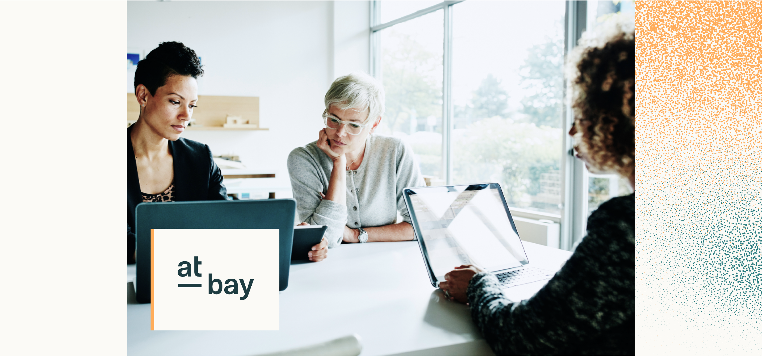 Three women sit at a table with laptops and a tablet, engaged in a discussion in a bright, modern office.