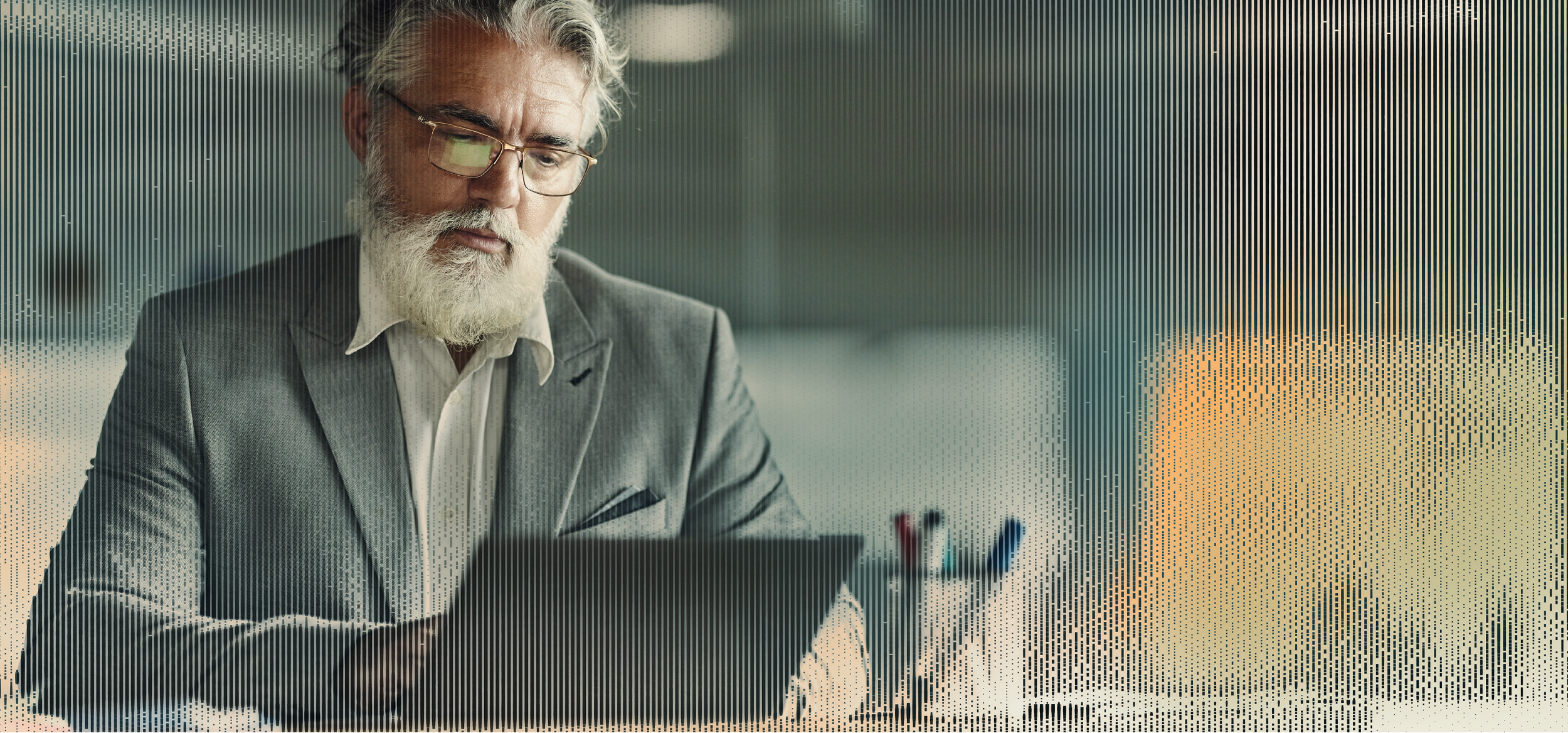 A senior man in a suit and glasses works on a laptop at a desk with office supplies.