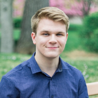 A young man with blonde hair wearing a blue shirt outdoors with greenery and pink blossoms in the background.