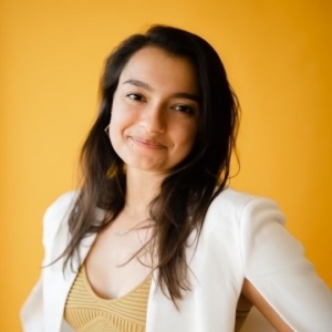 A young woman with long dark hair, smiling softly, wearing a white blazer over a beige top, against a yellow background.