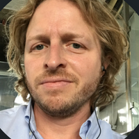 A man with wavy, shoulder-length hair and a light beard, wearing a blue shirt and earphones, in an indoor setting.