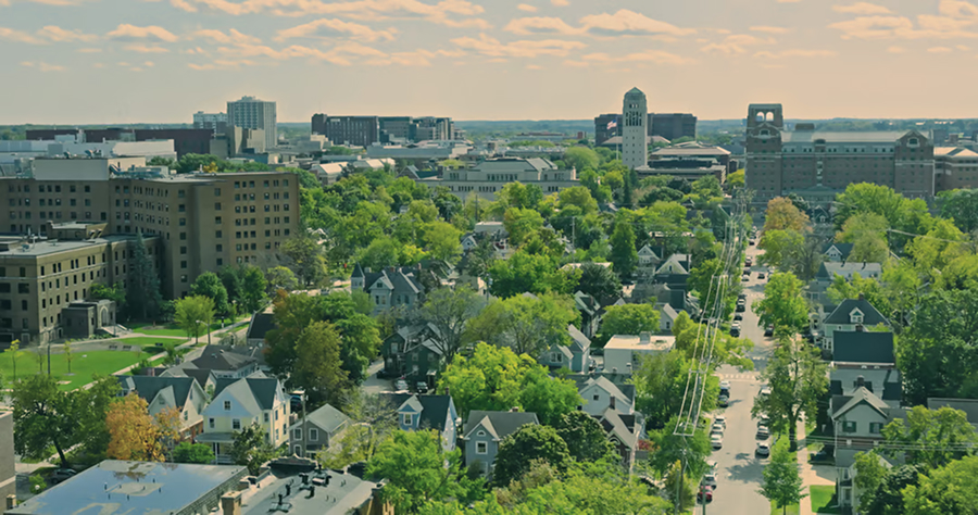 Aerial view of Ann Arbor, MI showing a mix of residential houses, green trees, and downtown buildings under a partly cloudy