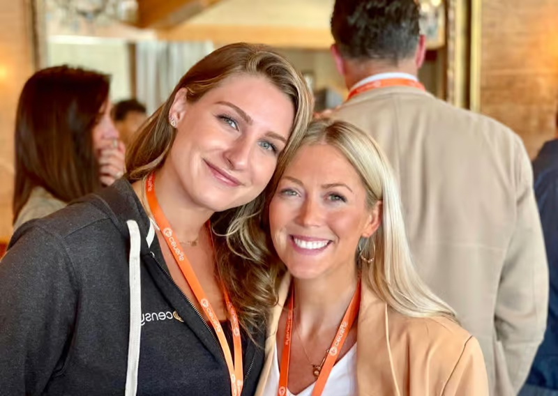 Two smiling women with orange lanyards pose together at a professional event, surrounded by other attendees in a warm,
