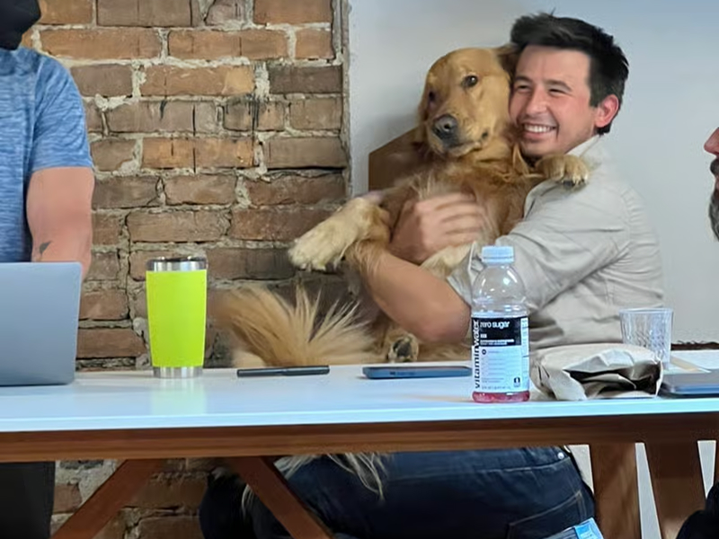 A man happily hugs a golden retriever while seated at a table with a brick wall background.
