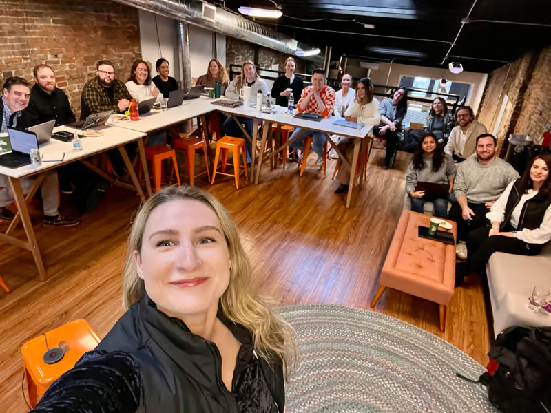 A woman takes a selfie in a room filled with diverse people seated at tables, smiling and working on laptops during a team