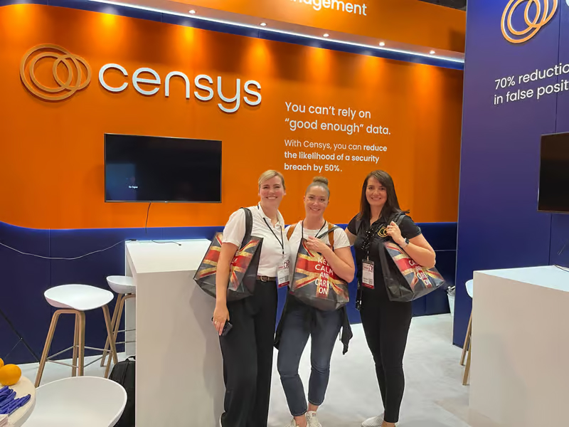 Three women stand together at a Censys booth, smiling and holding bags, in front of an orange and purple display.