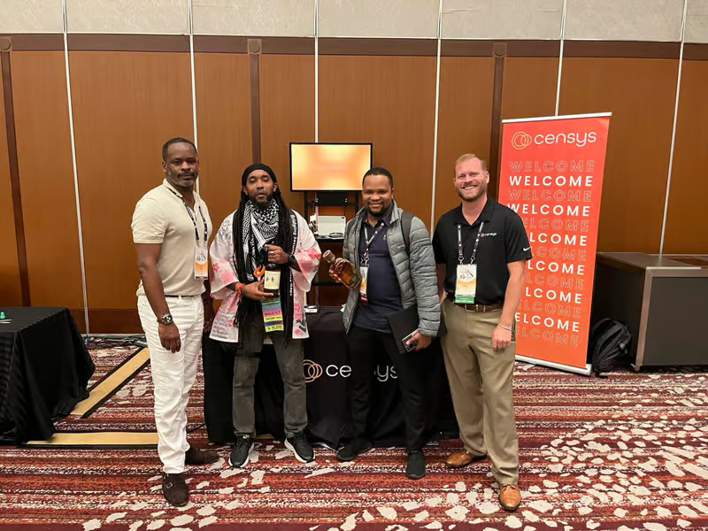 Four men stand together at a Censys event, smiling in front of a display table and a red "Welcome" banner.