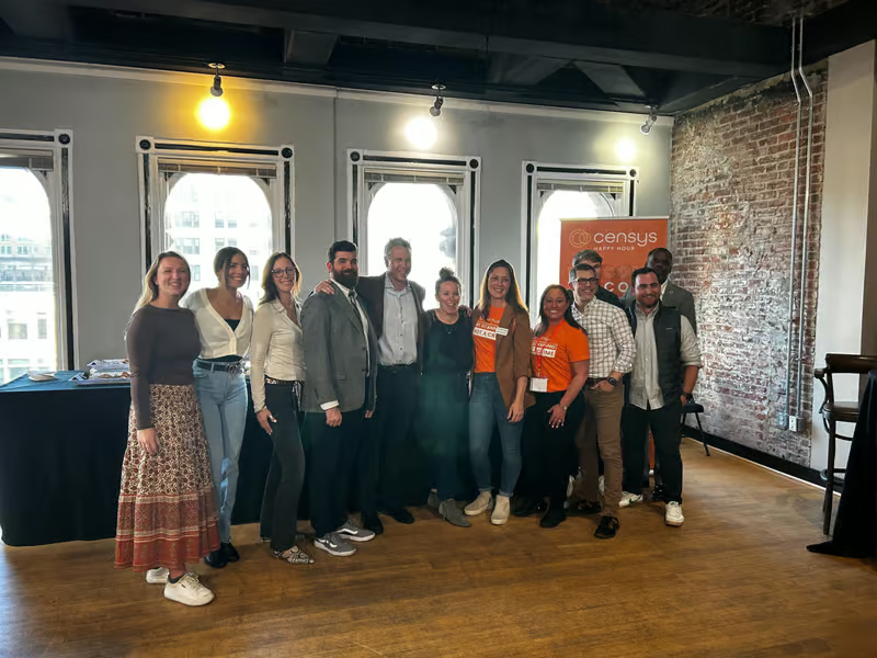 Group of diverse Censys team members smiling and posing together indoors with a Censys banner in the background.