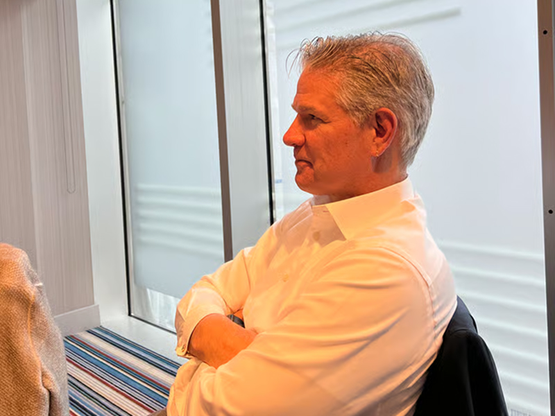 A mature man with gray hair in a white shirt sits with arms crossed, looking thoughtfully out a window.
