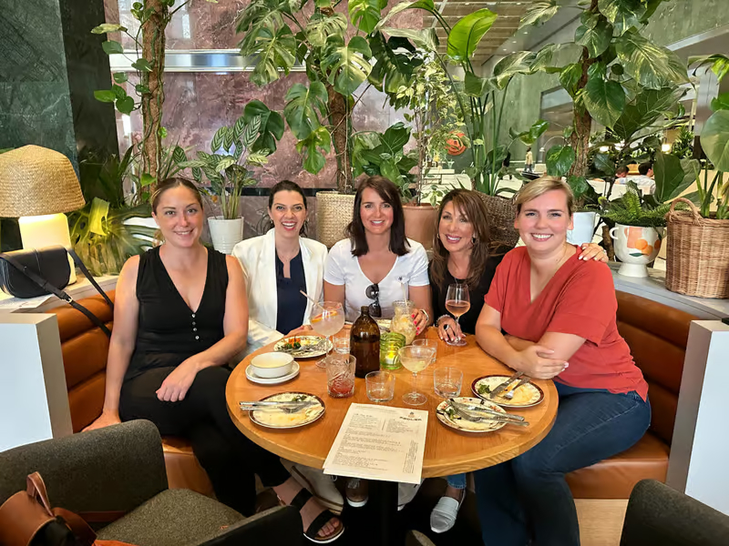 Five women sit together at a round table with drinks and plates, surrounded by lush green plants in a cozy restaurant