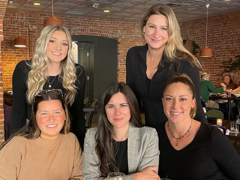 Five women gather at a restaurant, smiling and posing for a group photo in a cozy brick-walled setting.