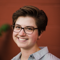 A young woman with short brown hair, glasses, and a light blue shirt, smiling against a wooden background.