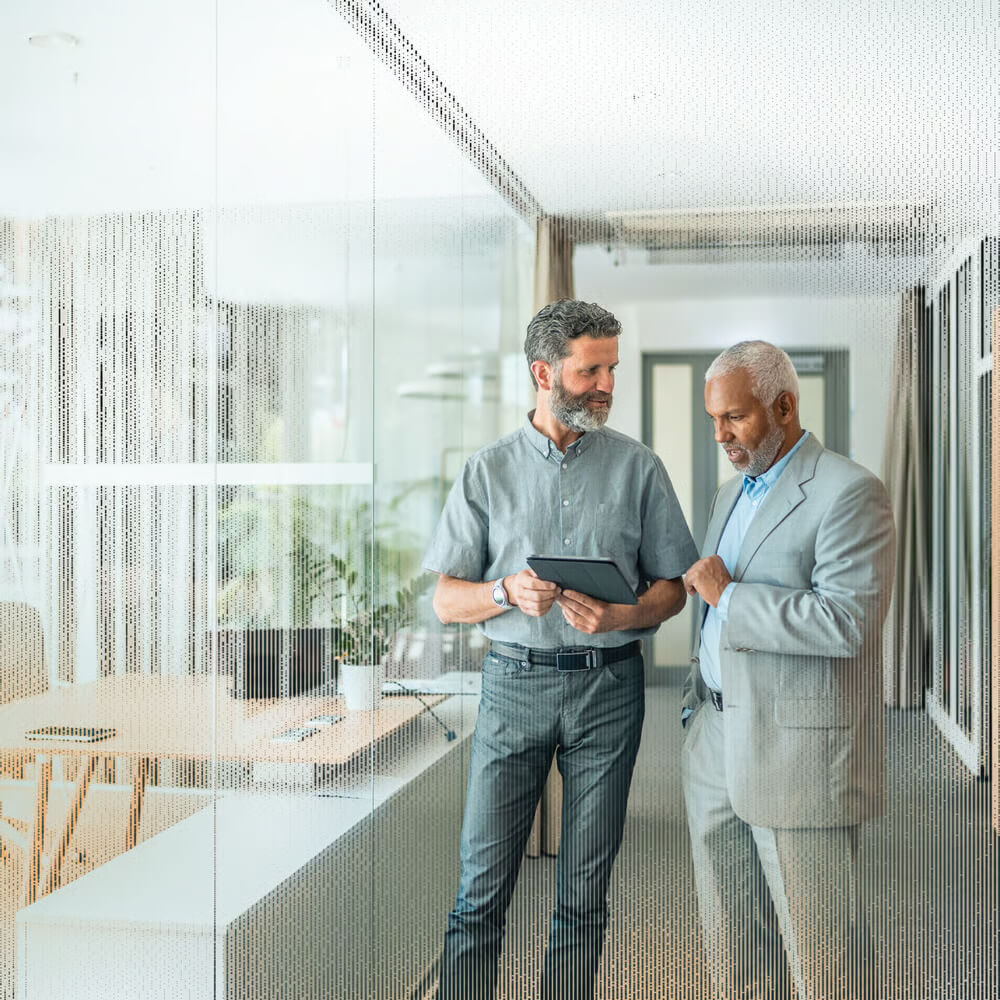 Two men in business attire discuss something on a tablet in a modern office with glass walls.
