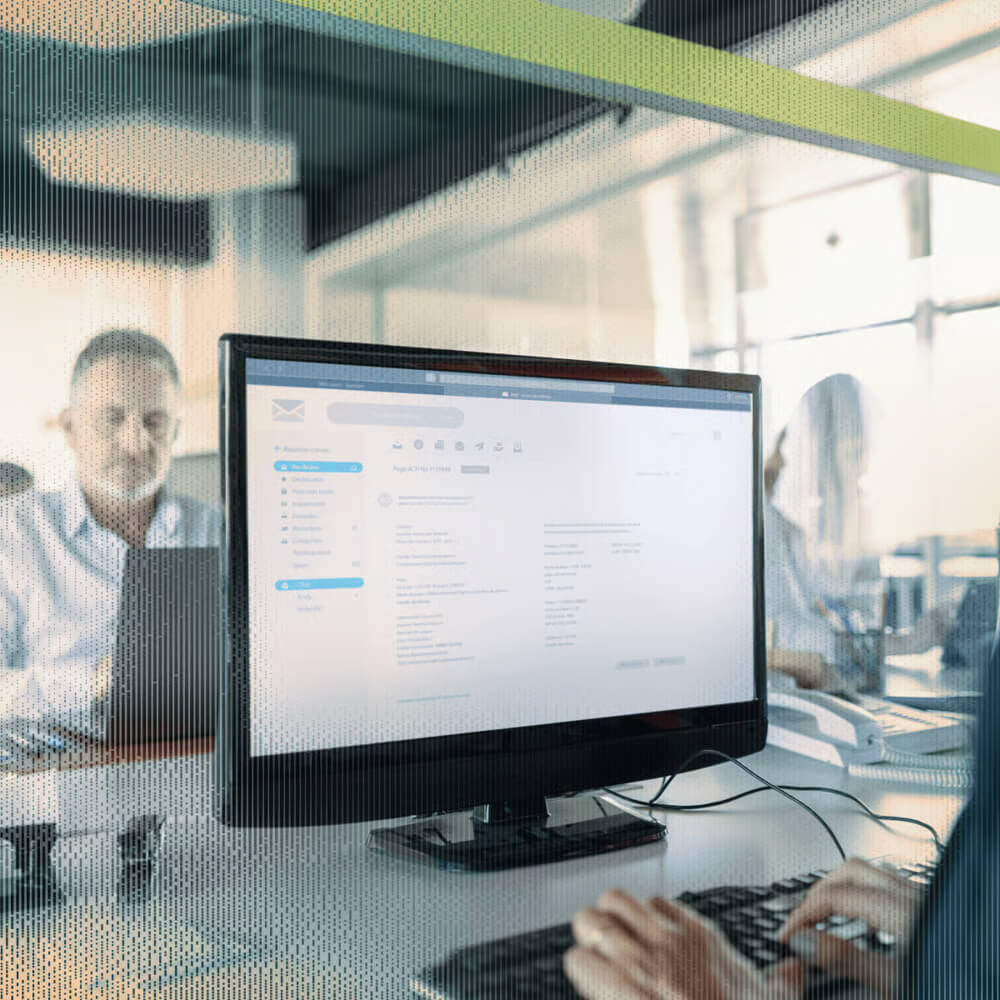 A person types on a keyboard while a monitor displays an email interface in an office setting.