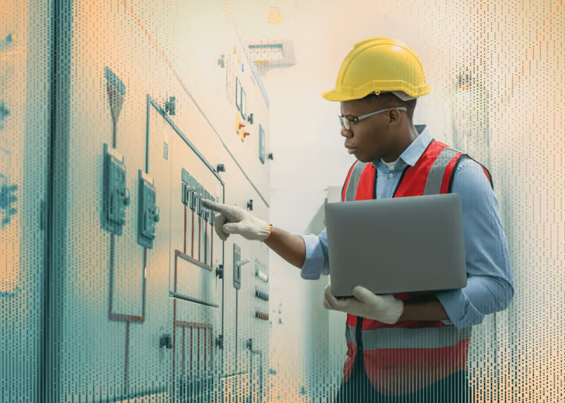 A technician in safety gear inspects electrical panels with a laptop in an energy utility facility.