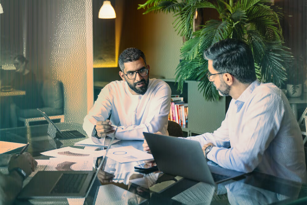 Two men in white shirts discuss cybersecurity strategies at a desk with laptops, papers, and a large plant in the background.