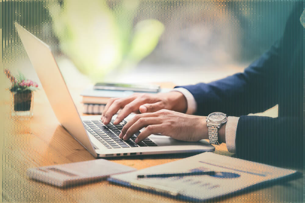 A person in a suit types on a laptop at a desk with documents, notebooks, and a potted plant, suggesting government use case