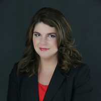A woman with shoulder-length wavy brown hair, wearing a black blazer and red top, poses against a dark background.