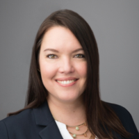 A woman with long dark hair, smiling, wearing a navy blazer and a gold necklace against a gray background.