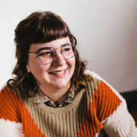 A smiling woman with dark hair, glasses, and a colorful sweater against a plain background.