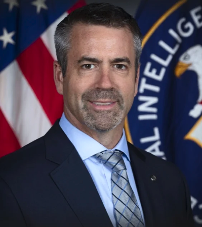 Andrew Boyd in a suit, standing in front of a U.S. flag and an intelligence agency emblem, smiling confidently.