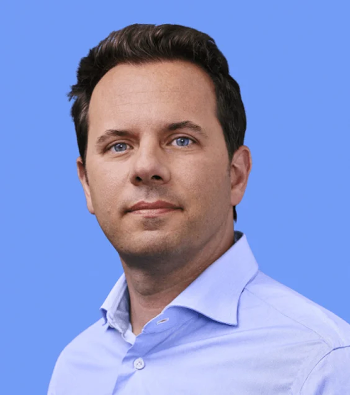 Karim Faris, a man with dark hair and blue eyes, wearing a light blue shirt, poses against a blue background.