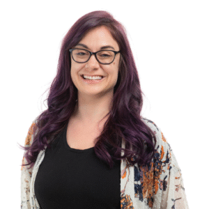 A woman with long, wavy purple hair, wearing glasses and a black top, smiling against a white background.