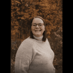 A smiling woman with glasses and shoulder-length hair wearing a beige sweater outdoors with autumn foliage in the background.