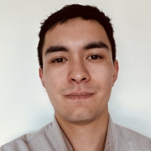 A young man with short dark hair and light skin, wearing a light-colored shirt, smiles softly against a plain background.