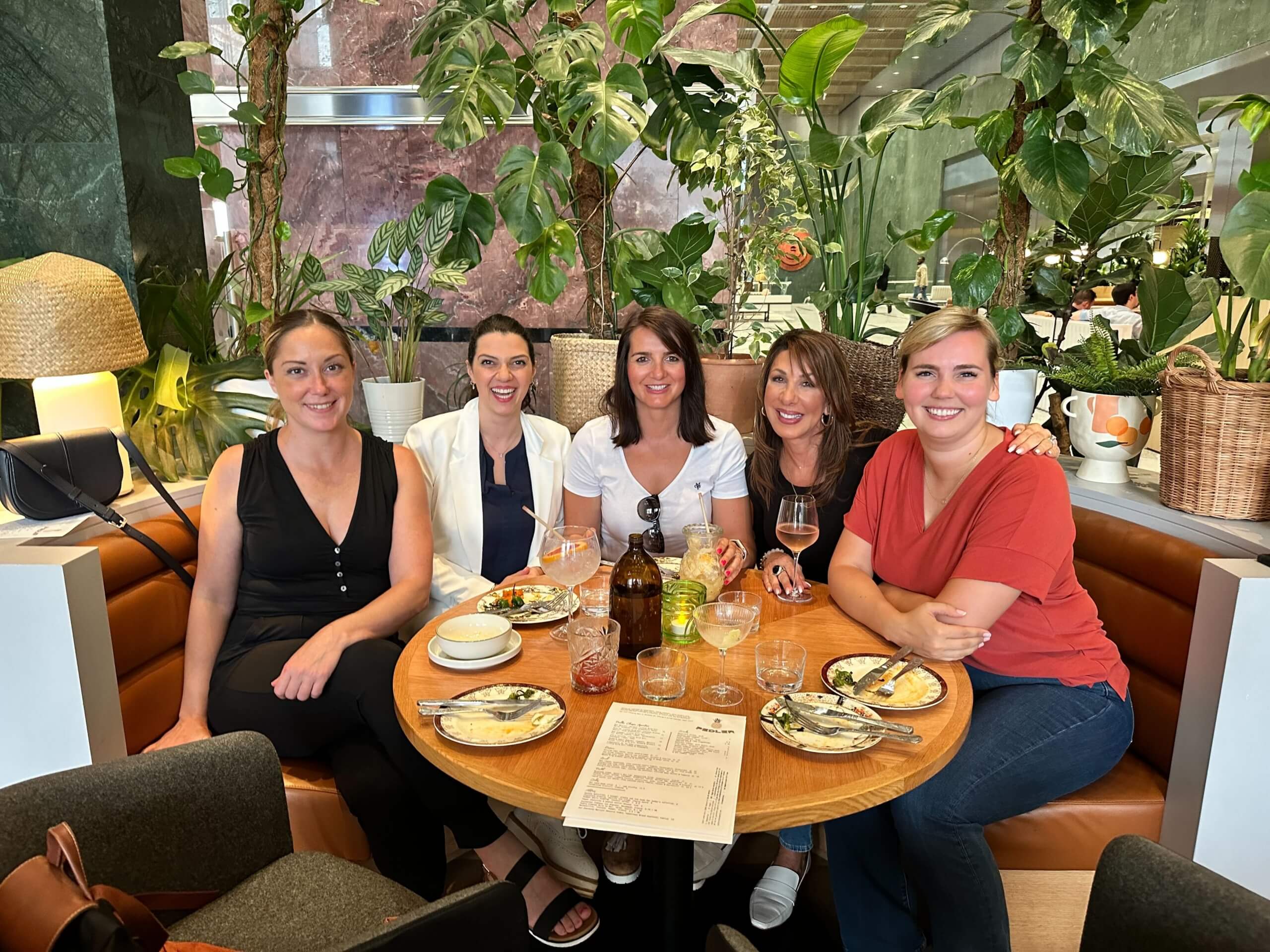 Five smiling Censys team members enjoying a meal together at a plant-filled restaurant with drinks and plates on the table.