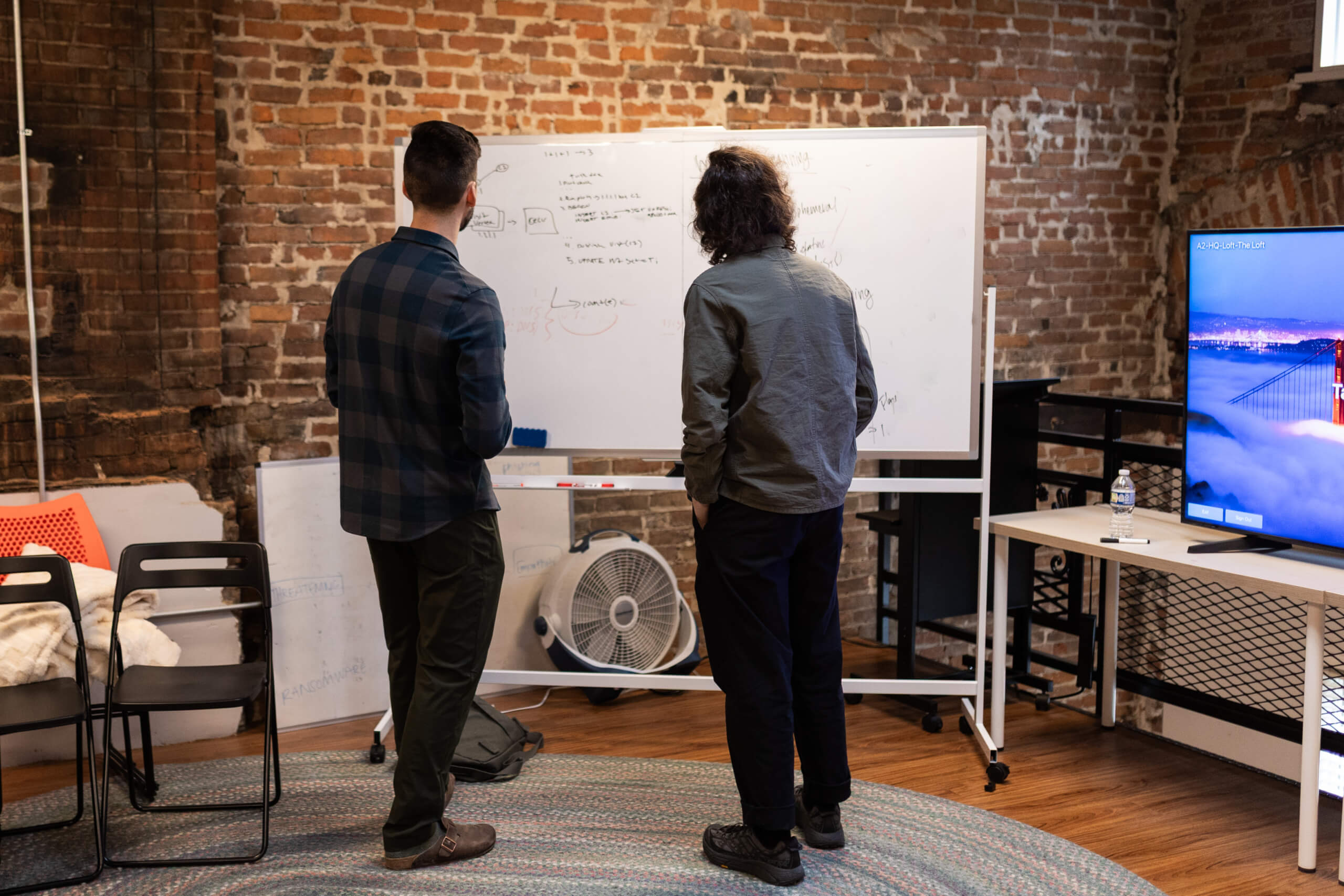 Two colleagues stand facing a whiteboard with notes in a brick-walled office space with a monitor nearby.