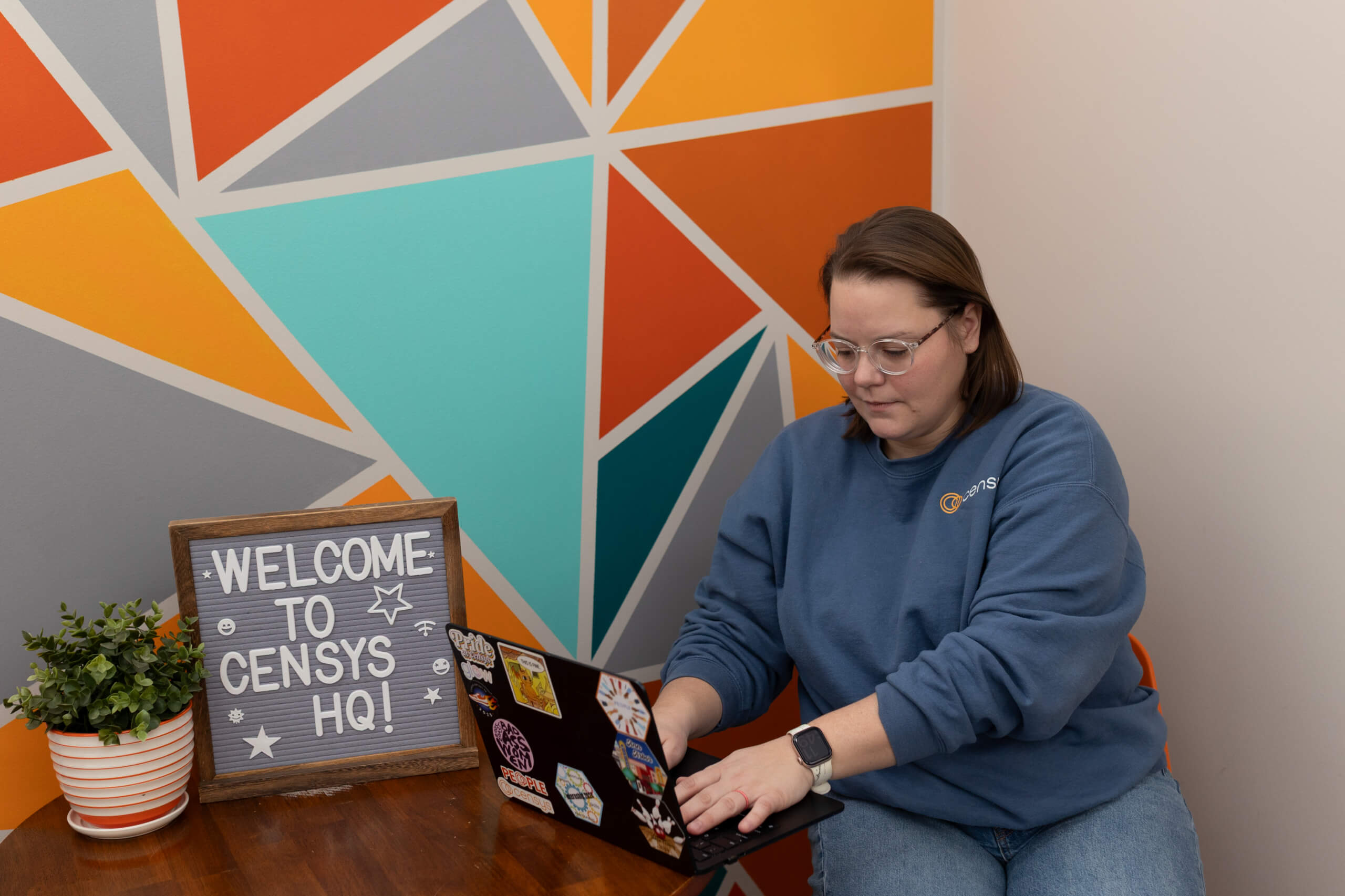 Censys employee in a branded sweatshirt works on a sticker-covered laptop beside a "Welcome to Censys HQ!" sign.