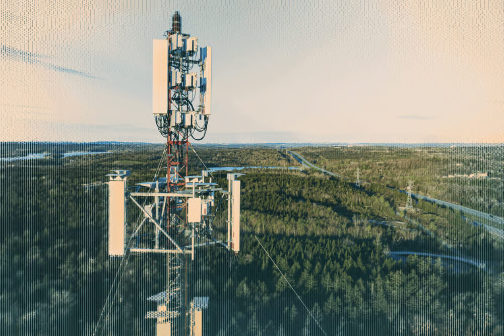 A tall communication tower with multiple antennas stands above a green forested landscape under a clear sky.