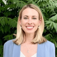A smiling woman with shoulder-length blonde hair, wearing a light blue blazer and a necklace, against a leafy green