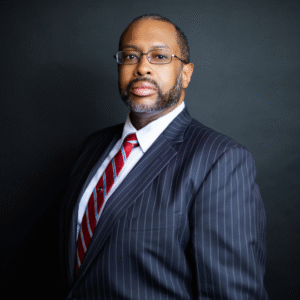 A confident man with glasses, a beard, and a pinstripe suit, wearing a red and white striped tie against a dark background.