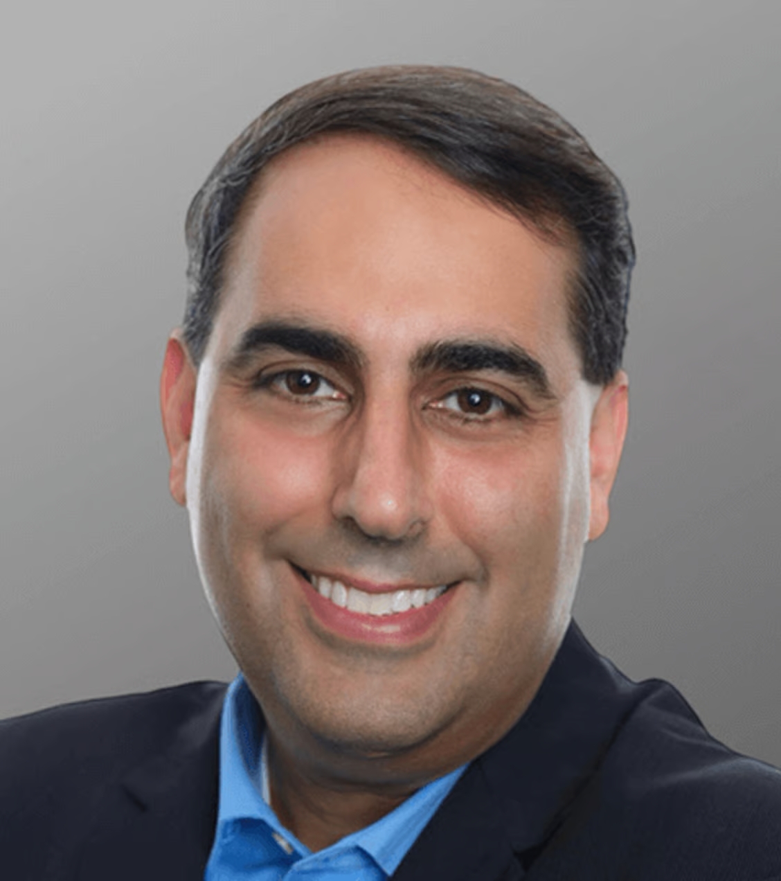 Headshot of a smiling man with dark hair, wearing a dark suit and blue shirt, against a gray background.