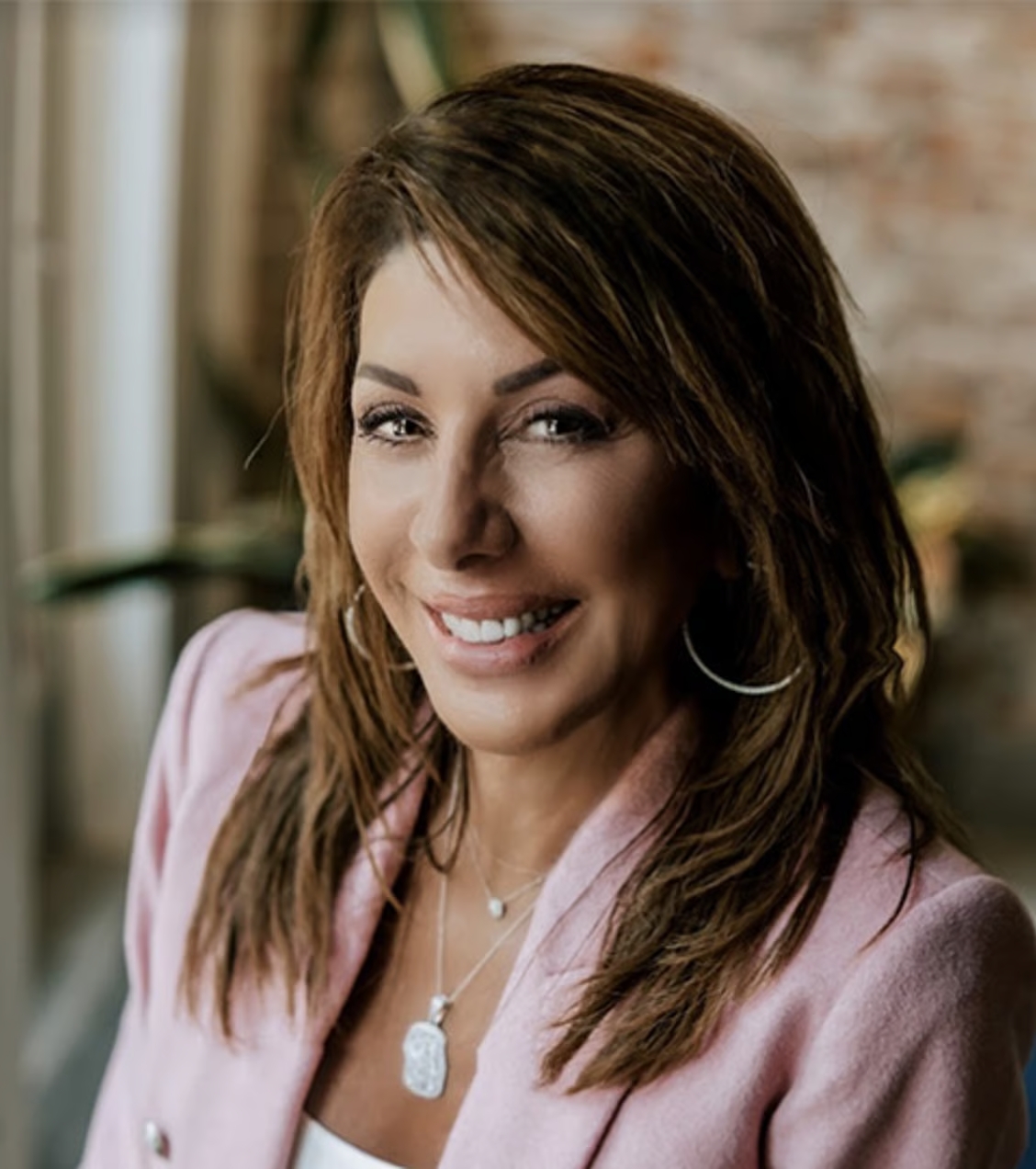 Sarah Ashburn with shoulder-length brown hair, wearing hoop earrings and a pink blazer, smiling warmly.
