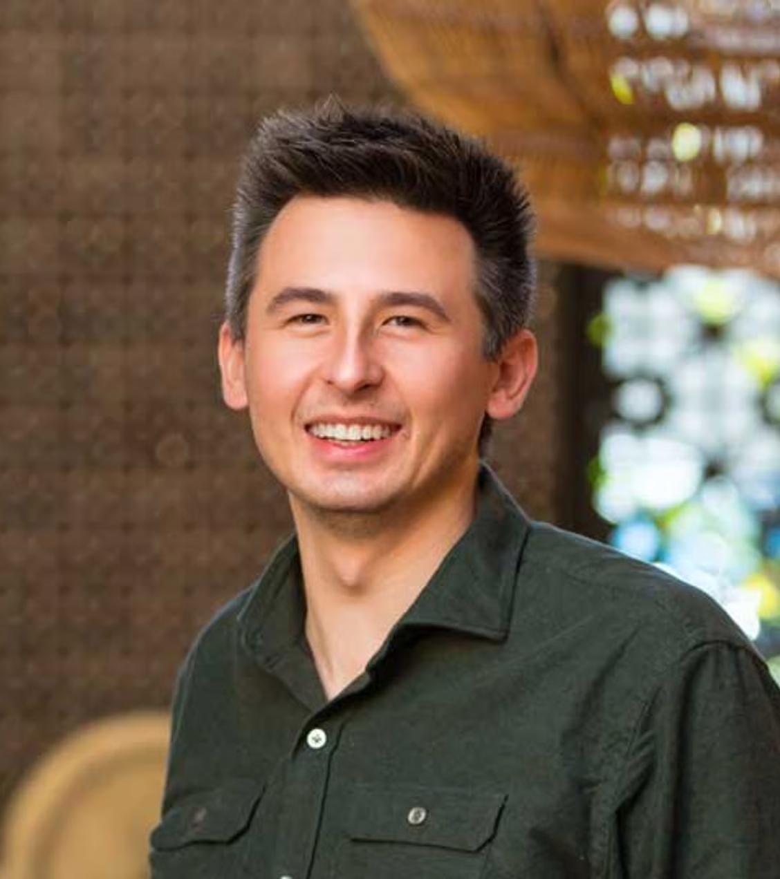 Headshot of a smiling man with dark, styled hair wearing a dark green shirt in a natural, outdoor setting.